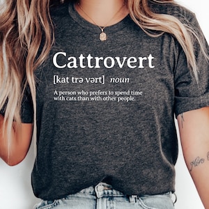 Op de afbeelding: Een donkergrijs T-shirt met het woord "Cattrovert" gedefinieerd als "een persoon die liever tijd doorbrengt met katten dan met andere mensen". Het shirt wordt gedragen door een persoon met een tatoeage op zijn arm die "glore Océans" zegt.