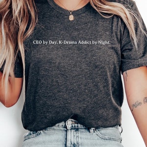 Puede incluir: Una camiseta gris oscuro con el texto "CEO by Day, K-Drama Addict by Night." impreso en blanco.