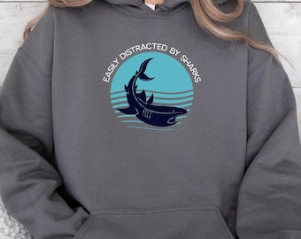 Può includere: Una felpa con cappuccio grigio scuro con un grafico di uno squalo in un cerchio blu. Il testo "Easily Distracted by Sharks" è scritto attorno al cerchio.