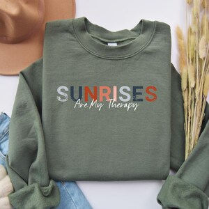 May include: A green sweatshirt with the text "SUNRISES Are My Therapy" in colourful block letters.