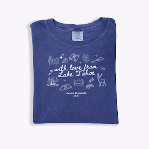 May include: A blue Comfort Colors t-shirt with a white graphic design. The design includes trees, binoculars, a campfire, a tent, and the text "with love from Lake Tahoe" and "Sarah's Bachelorette 2025".
