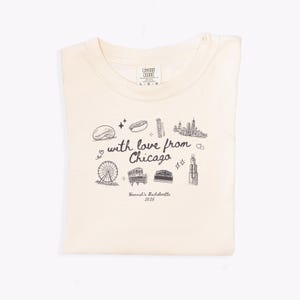 May include: Cream-colored t-shirt with a black graphic design featuring Chicago landmarks and the text "with love from Chicago." The shirt also includes the text "Hannah's Bachelorette 2025."