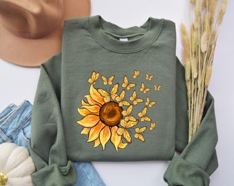 Könnte beinhalten: Ein grünes Sweatshirt mit einem aufgedruckten Sonnenblumen- und Schmetterlingsmotiv auf der Vorderseite. Die Sonnenblume ist gelb mit einer braunen Mitte und die Schmetterlinge sind gelb.