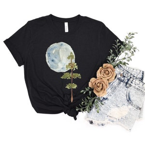 Puede incluir: Una camiseta negra con un dise&ntilde;o de acuarela de una luna azul y un &aacute;rbol verde. La camiseta est&aacute; anudada en la parte inferior.