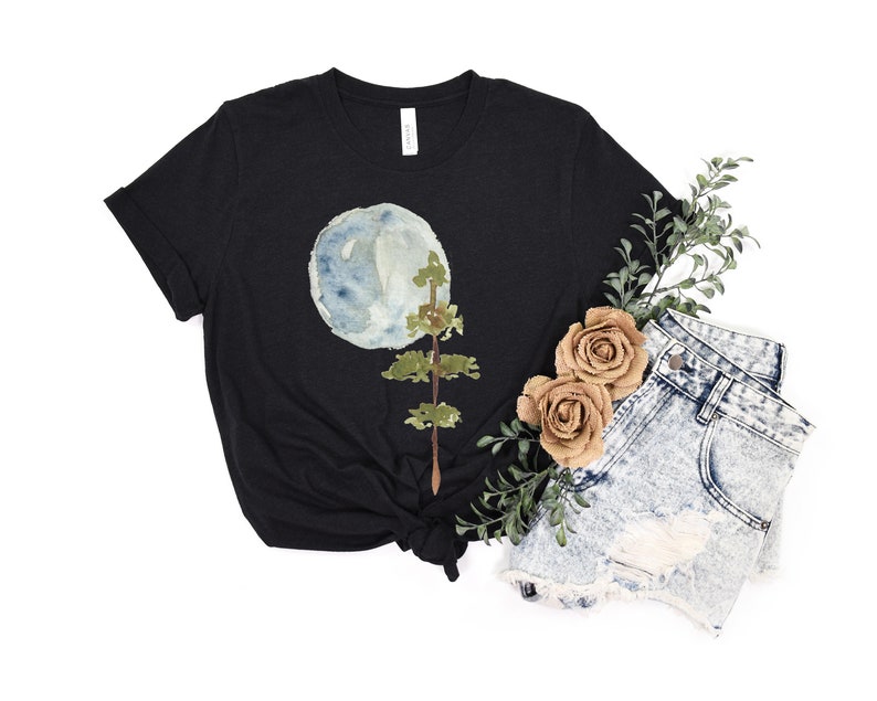Puede incluir: Una camiseta negra con un dise&ntilde;o de acuarela de una luna azul y un &aacute;rbol verde. La camiseta est&aacute; anudada en la parte inferior.