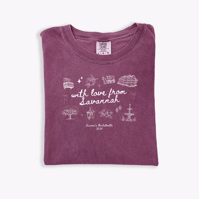 May include: A maroon Comfort Colors t-shirt with a crew neck. The shirt features a white graphic design with the text "with love from Savannah" and illustrations of landmarks. The text "Lauren's Bachelorette 2025" is also printed on the shirt.