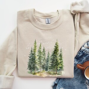 May include: A beige sweatshirt with a watercolor design of a forest scene. The design features a path leading into a grove of pine trees with a light dusting of snow.