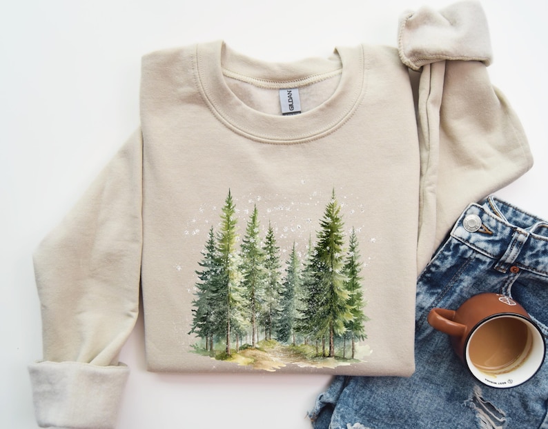 May include: A beige sweatshirt with a watercolor design of a forest scene. The design features a path leading into a grove of pine trees with a light dusting of snow.