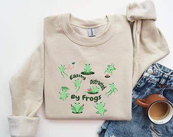 Puede incluir: Una sudadera beige con un gráfico de ranas verdes y el texto "Easily Distracted by Frogs".