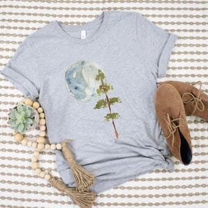 Puede incluir: Una camiseta gris con un dise&ntilde;o de acuarela de una luna y un &aacute;rbol. La camiseta est&aacute; sobre un fondo a rayas con una guirnalda de cuentas de madera y botas de gamuza marr&oacute;n.