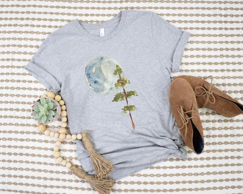 Puede incluir: Una camiseta gris con un dise&ntilde;o de acuarela de una luna y un &aacute;rbol. La camiseta est&aacute; sobre un fondo a rayas con una guirnalda de cuentas de madera y botas de gamuza marr&oacute;n.