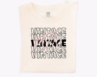 May include: A beige t-shirt with the text "Vintage 1985" printed in black and pink. The text is stacked and repeated multiple times.