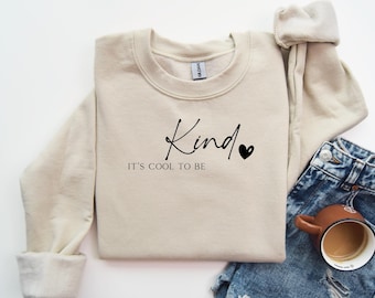 Könnte beinhalten: Ein beiger Pullover mit dem Text "Kind, It's cool to be" in schwarzer Schrift und einem kleinen schwarzen Herz.