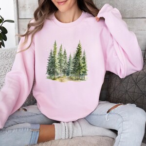 May include: A pink sweatshirt with a watercolor illustration of a forest scene. The trees are green and the background is a light blue.