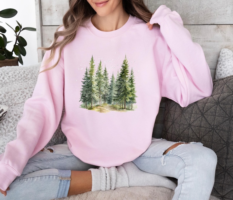 May include: A pink sweatshirt with a watercolor illustration of a forest scene. The trees are green and the background is a light blue.