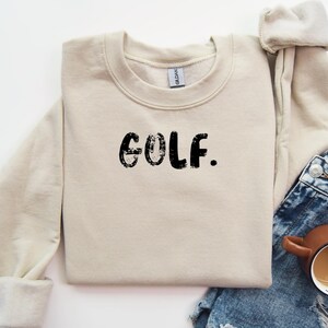 May include: A beige sweatshirt with the word "GOLF" printed in black on the front. The sweatshirt is folded and laid on a white surface.