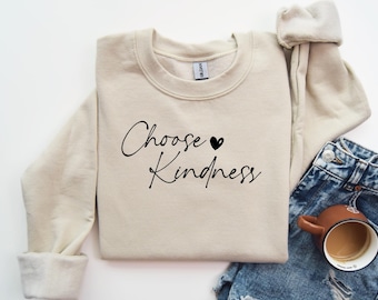 Könnte beinhalten: Ein beiger Pullover mit dem Text "Choose Kindness" in schwarzer Schrift mit einem kleinen schwarzen Herzen über dem Wort "Kindness".