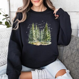 May include: A black sweatshirt with a watercolor illustration of a snowy forest scene. The trees are green and the snow is white. The sweatshirt is a comfortable and stylish choice for winter.