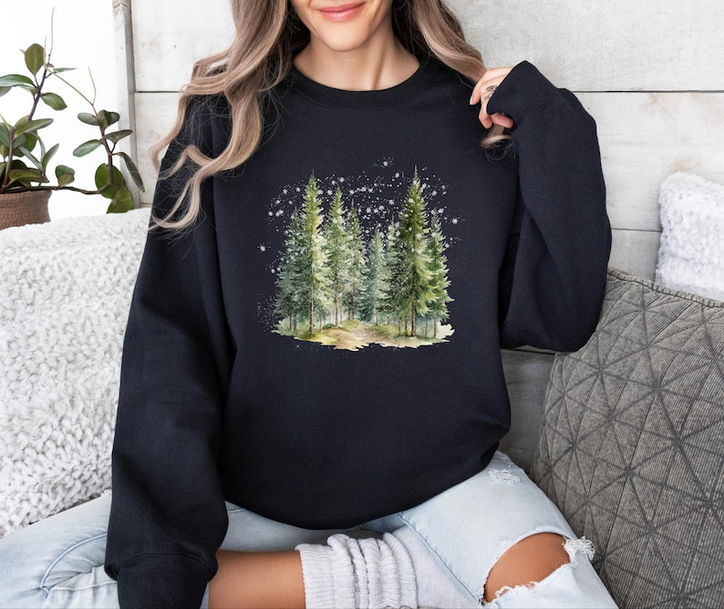 May include: A black sweatshirt with a watercolor illustration of a snowy forest scene. The trees are green and the snow is white. The sweatshirt is a comfortable and stylish choice for winter.