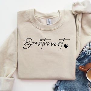 Può includere: Una felpa beige con il testo "Booktrovert" in scrittura nera con un piccolo cuore nero.