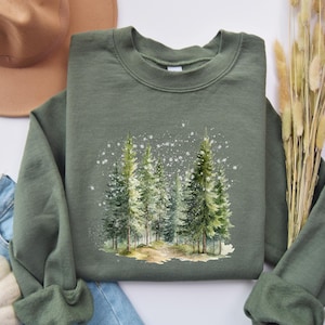 May include: A green sweatshirt with a watercolor design of a snowy forest. The design features several pine trees with white snowflakes falling around them.