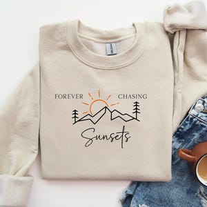 May include: A beige sweatshirt with the text "Forever Chasing Sunsets" and a graphic of a sun, mountains, and trees.