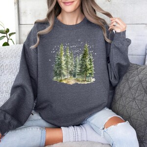 May include: A dark gray sweatshirt with a watercolor illustration of a snowy forest scene. The illustration features a group of pine trees with a path leading through them.