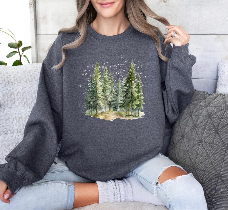 May include: A dark gray sweatshirt with a watercolor illustration of a snowy forest scene. The illustration features a group of pine trees with a path leading through them.