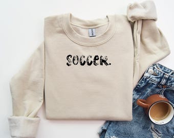 Puede incluir: Una sudadera beige de cuello redondo con la palabra "soccer" impresa en negro en la parte delantera.