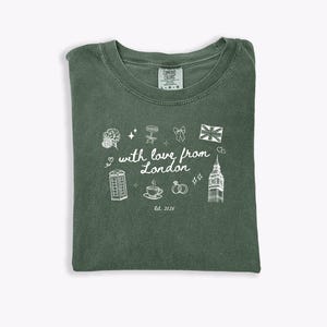 May include: Green Comfort Colors t-shirt with a white graphic design. The design includes a bouquet, stars, a bow, a Union Jack, a phone booth, a cup of tea, wedding rings, Big Ben, and the text "with love from London" and "Est. 2025".