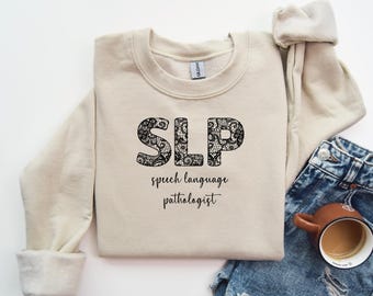 May include: A beige sweatshirt with the text "SLP speech language pathologist" in black lettering. The letters are decorated with a black lace pattern.