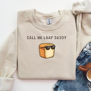 May include: A beige sweatshirt with the text "Call me Loaf Daddy" and a cartoon image of a loaf of bread wearing sunglasses.