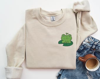 Puede incluir: Una sudadera beige con una rana verde bordada en la parte delantera. La rana está sentada en un nenúfar verde con un pequeño brote verde que le crece de la cabeza.