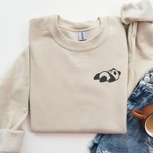 May include: A beige sweatshirt with a small black and white panda bear design on the front. The sweatshirt is folded and laid on a white surface with a pair of blue denim shorts and a brown coffee mug.