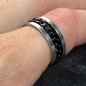 May include: A silver ring featuring a central band of black chain links. The ring is worn on a finger, showcasing the intricate chain pattern. The ring has a polished silver finish and a contemporary design.