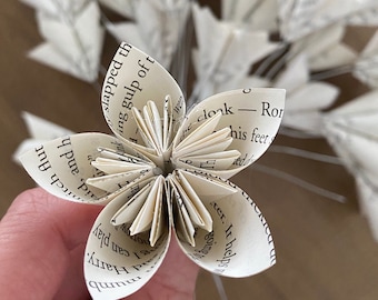 Peut inclure: Gros plan sur une fleur en papier faite à partir de pages de livre. Les pétales blancs présentent du texte imprimé. La fleur est tenue, avec d'autres fleurs similaires floues en arrière-plan. La fleur est fixée à une tige métallique.