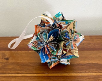 May include: A spherical ornament constructed from folded paper map segments featuring various colours and printed text. The structure consists of multiple petal-like sections arranged in a geometric pattern, with a white ribbon loop attached for hanging.