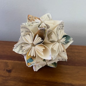 Peut inclure: Une boule de fleurs en papier blanc réalisée à partir de pages d'un livre. Les pages comportent du texte et des illustrations. La boule est composée de nombreuses fleurs individuelles, chacune ayant cinq pétales.