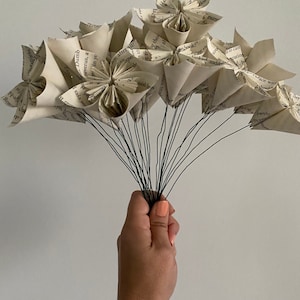 May include: A bouquet of paper flowers made from book pages. The flowers are white and have a vintage look. The stems are made of wire and are held together by a hand.