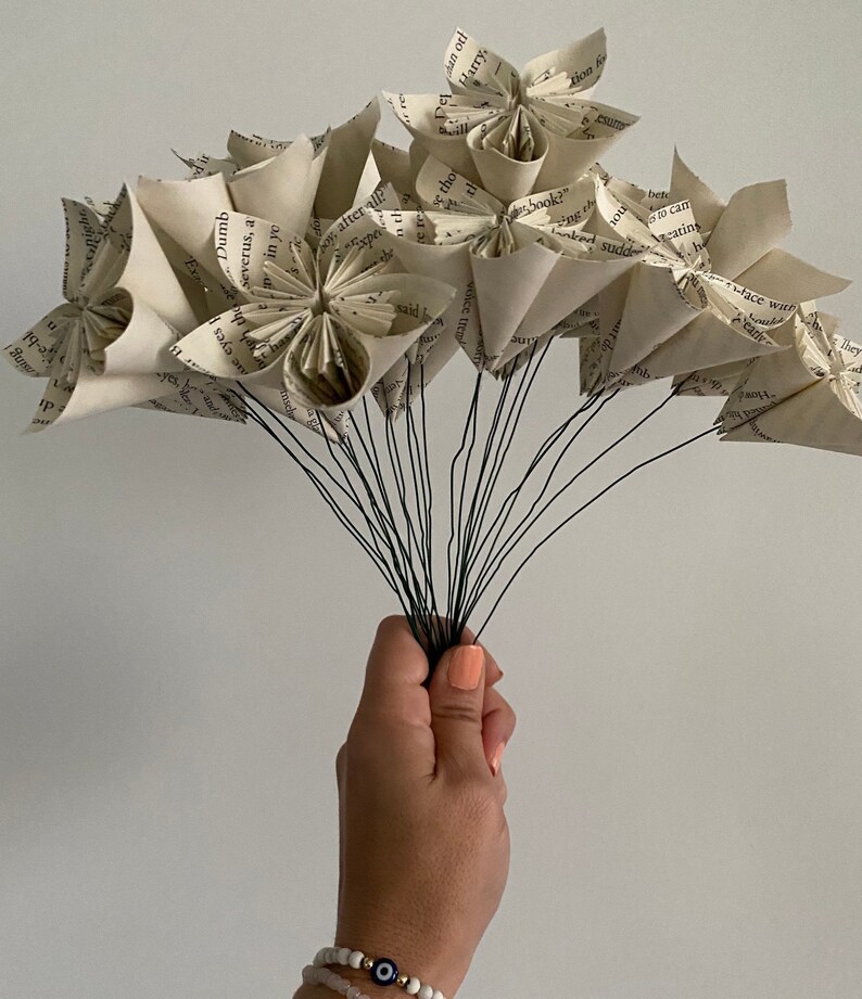May include: A bouquet of paper flowers made from book pages. The flowers are white and have a vintage look. The stems are made of wire and are held together by a hand.