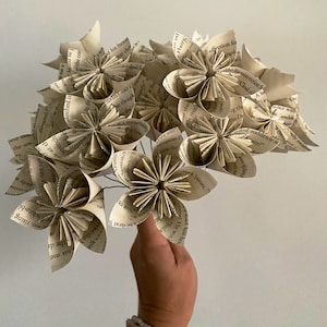 May include: A bouquet of paper flowers made from the pages of old books. The flowers are white and cream colored and have a vintage aesthetic.