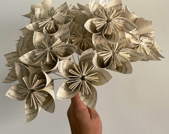 以下が含まれることがあります： 古い本のページで作られた紙の花のブーケ。花は白とクリーム色で、ヴィンテージな雰囲気です。