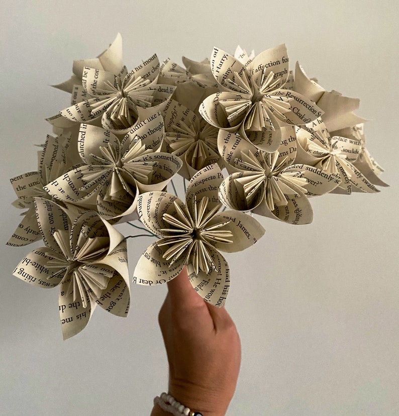 May include: A bouquet of paper flowers made from the pages of old books. The flowers are white and cream colored and have a vintage aesthetic.