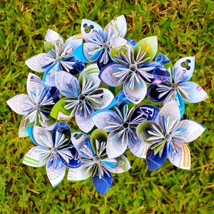 May include: A bouquet of handcrafted paper flowers made from recycled magazine pages. Each flower features multi-colored petals with visible printed text, graphics, and photographs in shades of blue, green, white, and yellow, arranged in a circular cluster.