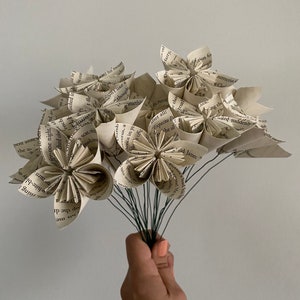 May include: A bouquet of paper flowers made from book pages. The flowers are white and cream colored and have a vintage look. The flowers are arranged in a bouquet and tied together with a brown ribbon.