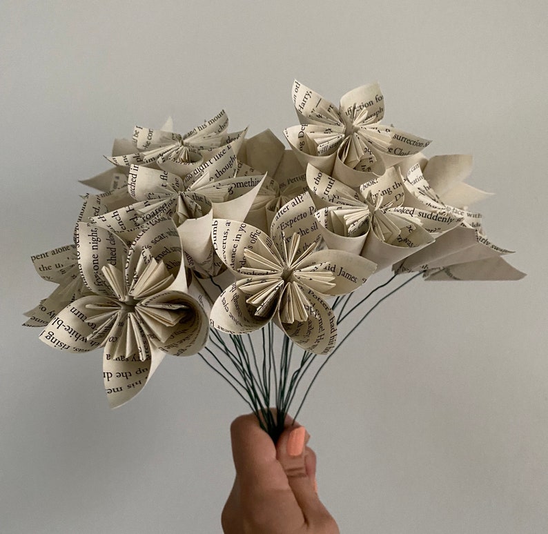 May include: A bouquet of paper flowers made from book pages. The flowers are white and cream colored and have a vintage look. The flowers are arranged in a bouquet and tied together with a brown ribbon.