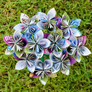 May include: A bouquet of handmade paper flowers with white, purple, blue, and green petals. The flowers are arranged in a circular shape, with green stems and leaves. The flowers are made from recycled paper.