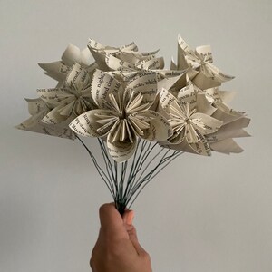 May include: A bouquet of paper flowers made from the pages of old books. The flowers are white and cream colored and have a rustic, vintage look. The stems are wired and ready to be arranged in a vase.