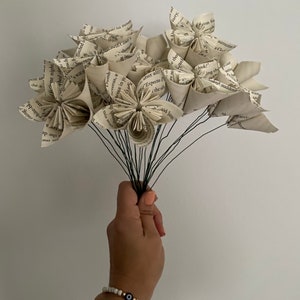 May include: A bouquet of paper flowers made from book pages. The flowers are white and have a delicate, vintage look. The stems are black wire.