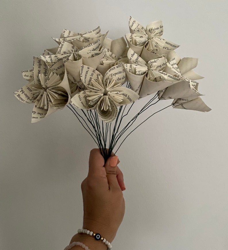 May include: A bouquet of paper flowers made from book pages. The flowers are white and have a delicate, vintage look. The stems are black wire.